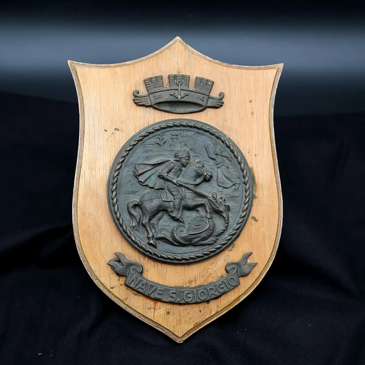 Italiensk marine-våbenskjold fra skibet S. Giorgio – givet ved officielt besøg på Holmen i sommeren 1966. Massiv messingmedaljon af Sankt Georg og dragen på træskjold med stærk patina.